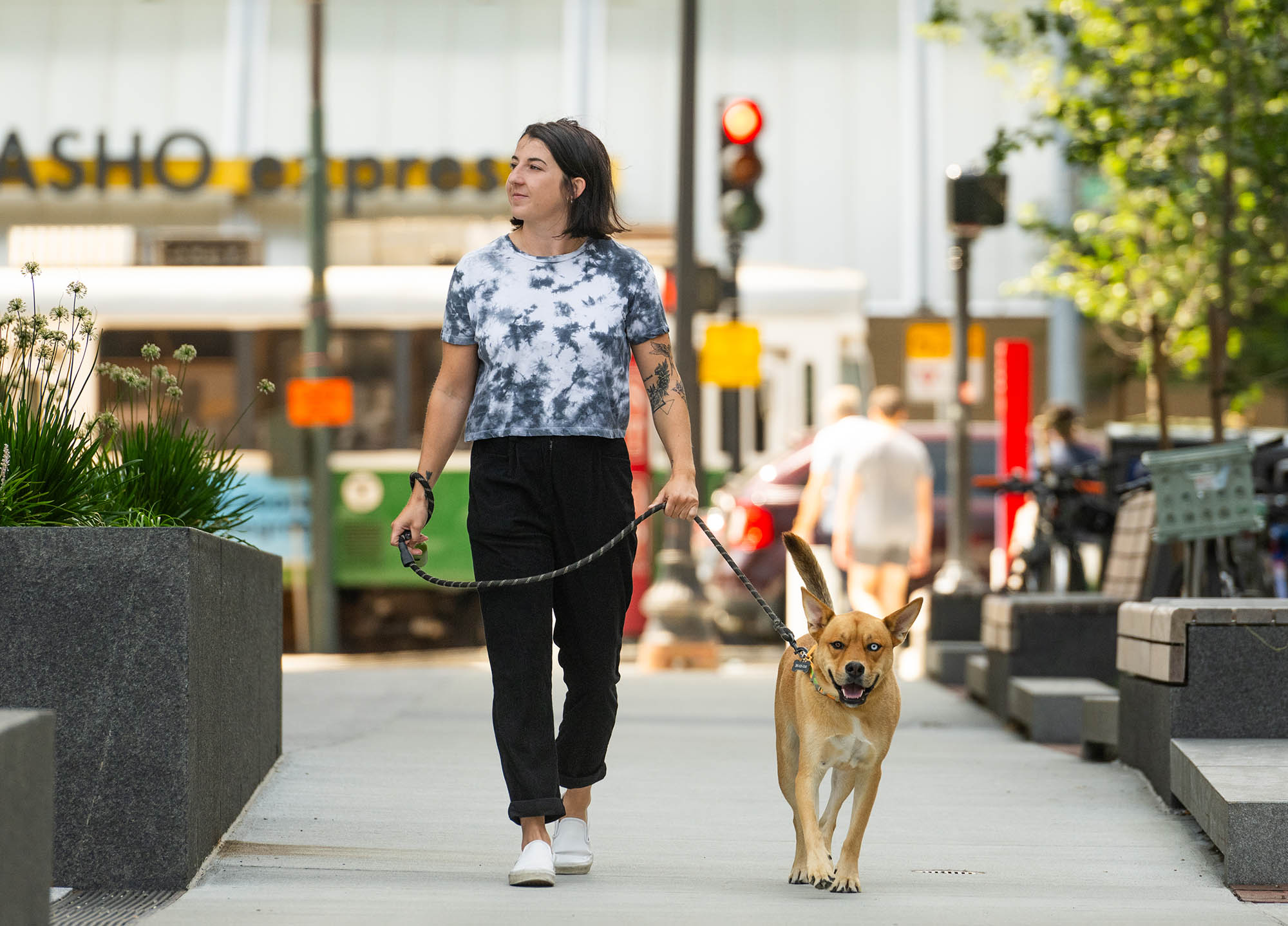 Photo: Chelsea Houlihan, a white woman wearing a grey and white tye-dye shirt and black pants, looks to the left as she holds the leash to her brown rescue mix dog. They both walk down a quiet street.
