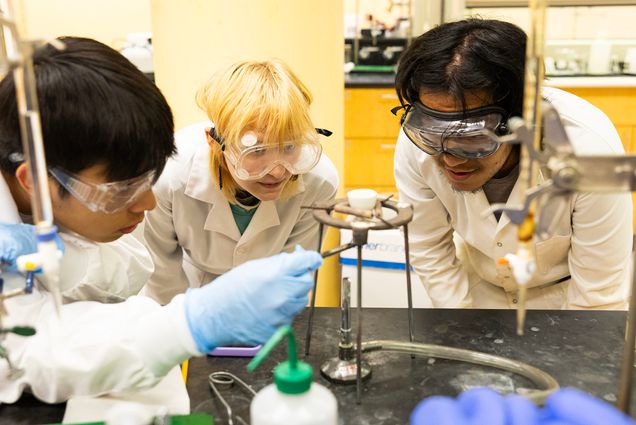 Photo: Three college students in white lab coats and goggles gather around a test tube burner apparatus and perform a chemistry experiment