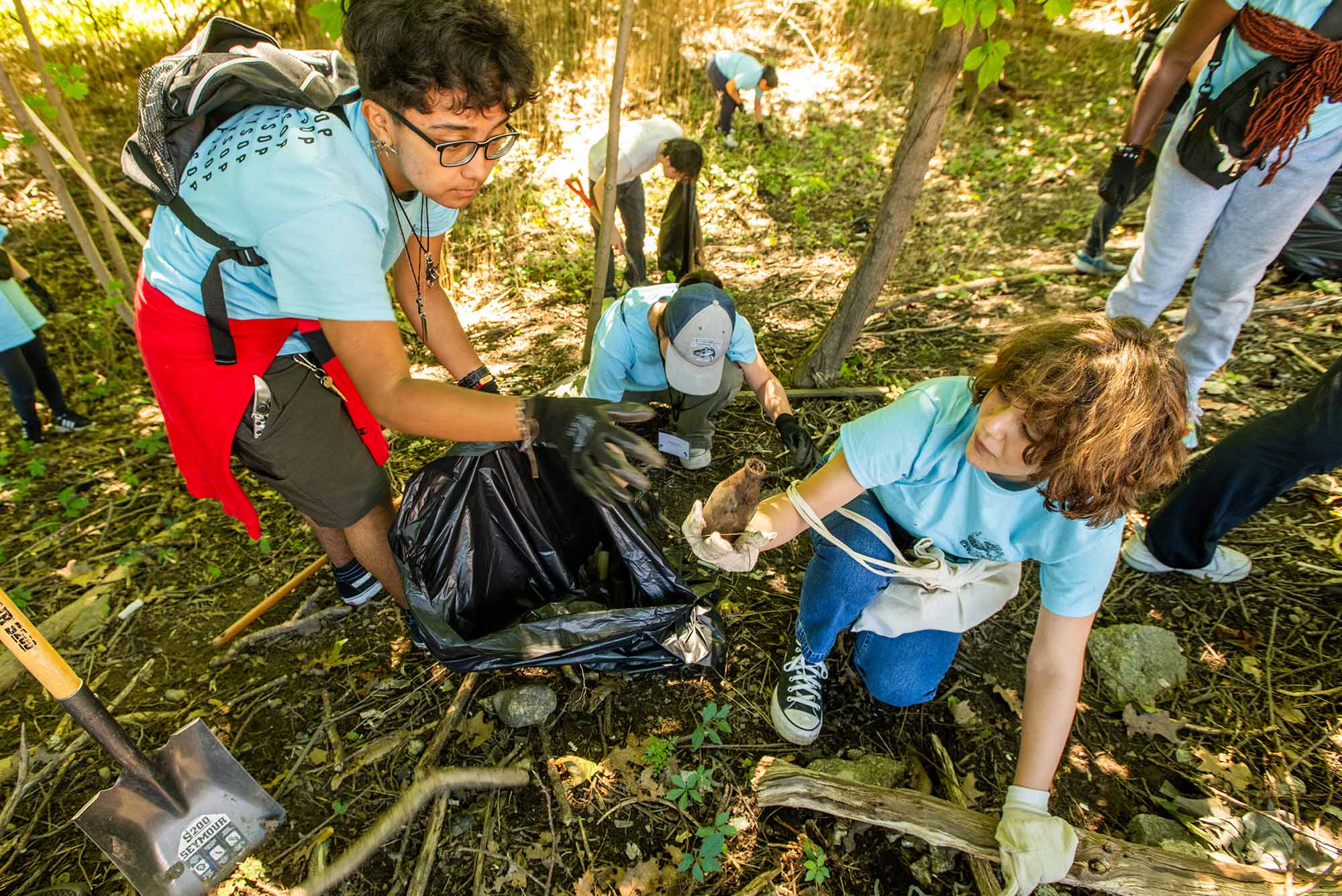 Photo: Two college students kneel and clean up trash in a forest-y, wooded area. They wear light blue shirts that read "FYSOP" and gardening gloves and hold black trash bags.