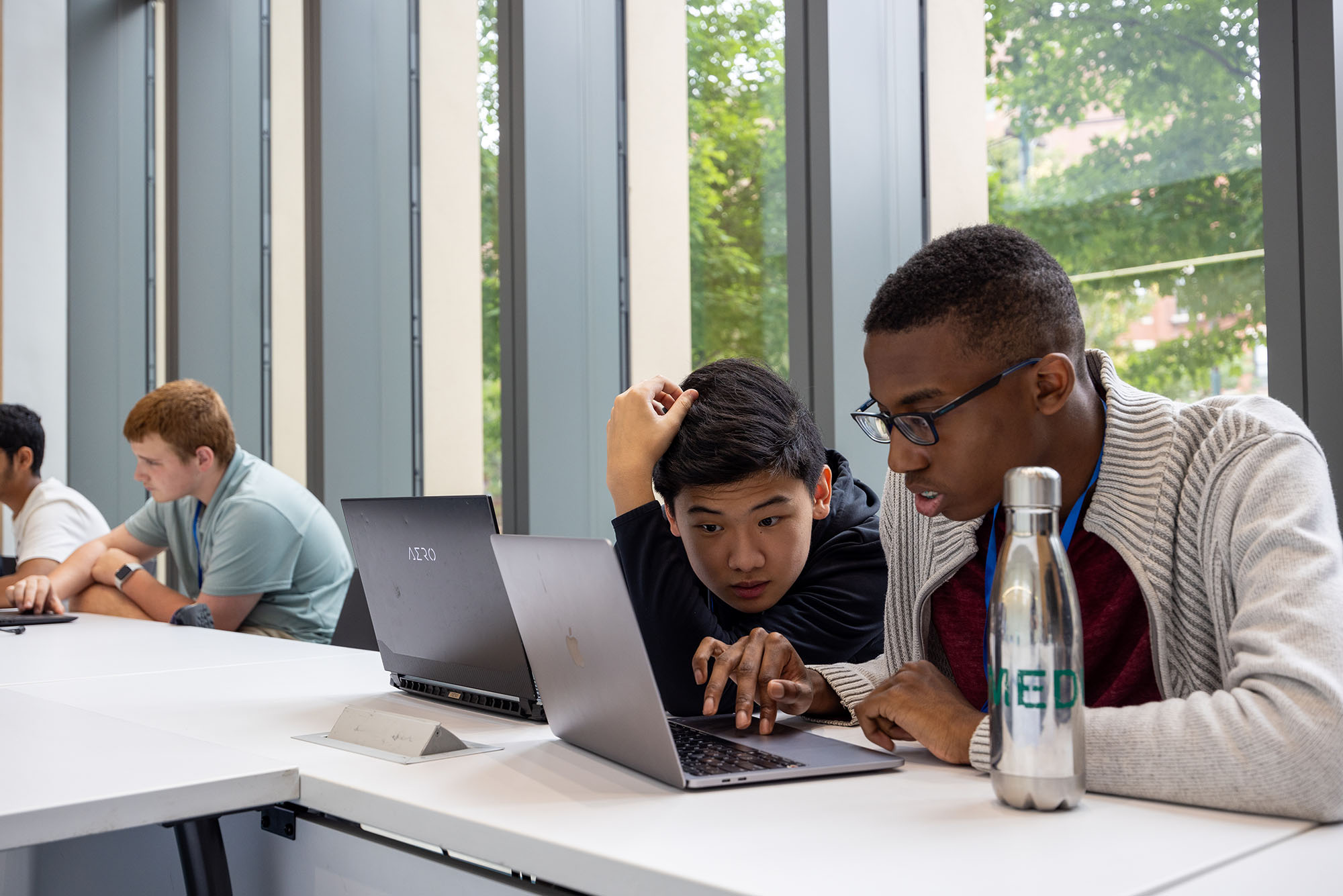 Photo: Eleventh graders David Luo (center), and Yareh Constant, both look closely at a laptop screen. A young tan boy (left) and a young Black boy look intently at a laptop screen as they sit at a long, white table. Other young teens can be seen looking at various laptop screens as they sit along the table as well.
