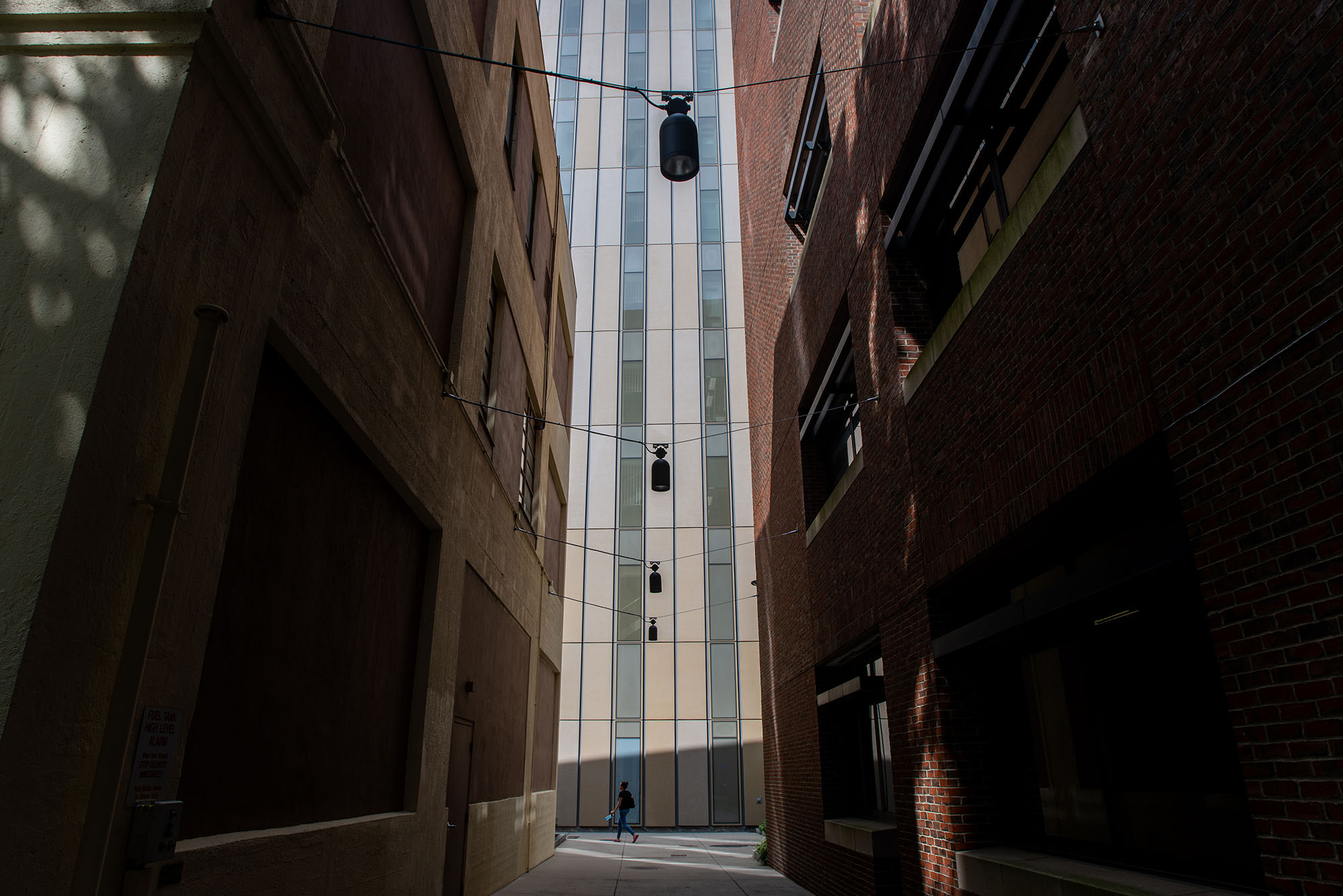 Photo: A pedestrian is shown passing by two buildings. Shot is taken in alley between two buildings and highlights the shadows and sun of the buildings.