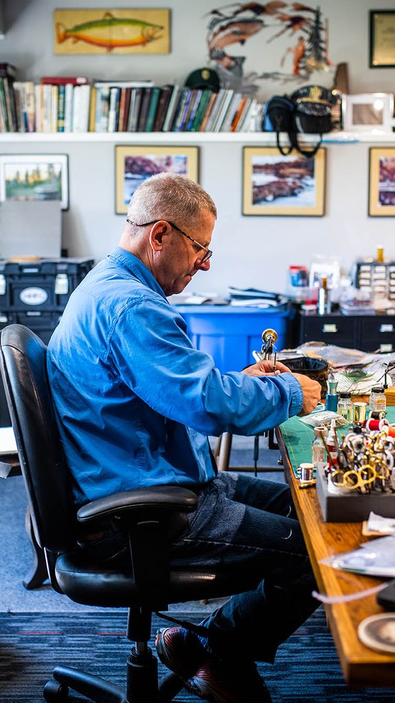 Photo: Scott Biron makes a fly at his New London, NH home. An older white man wearing glasses, a long-sleeved blue collared shirt, and jeans sits at a crowded desk and works on a fly. Fly tying materials and paraphernalia can be seen around the office.