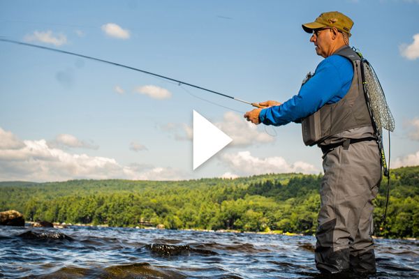 Photo: Scott Biron casts his fishing line out as he stands in knee-length water. An older white man wearing glasses, a long-sleeved blue collared shirt, and khaki water wader pants and hat, holds a fishing rod and casts out the line. He stands in water on a sunny day. A white triangle play button is shown in the center.