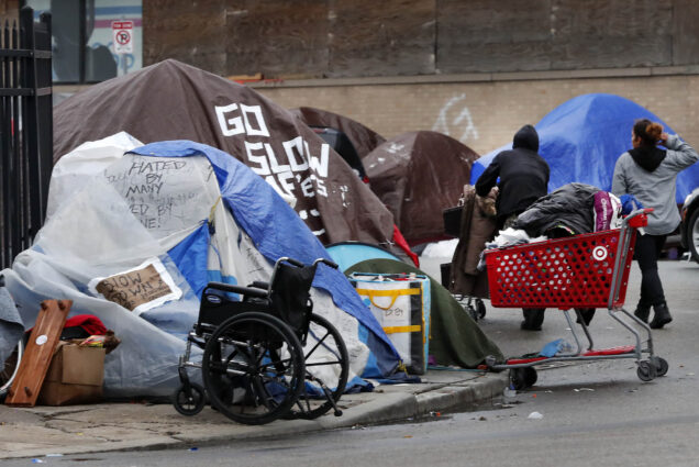 Photos: A group of mishapen tents are shown on a street of Boston. A lone red shopping cart filled with full trash bags and an old black wheelchair sit in front of the street. A few people can be seen amidst the tents.