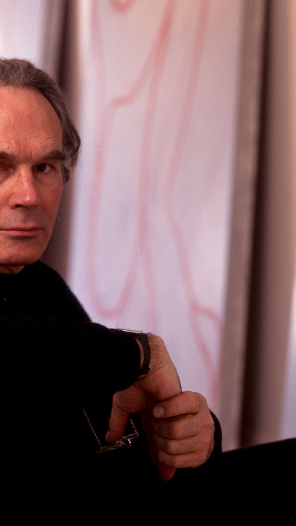 Photo: Portrait of Brice Marden, a white man with grey-brown hair and wearing black long sleeved shirt and pants, sits on a chair and looks over his shoulder at the camera. He sits in front of a wall full of large canvases featuring his paintings.