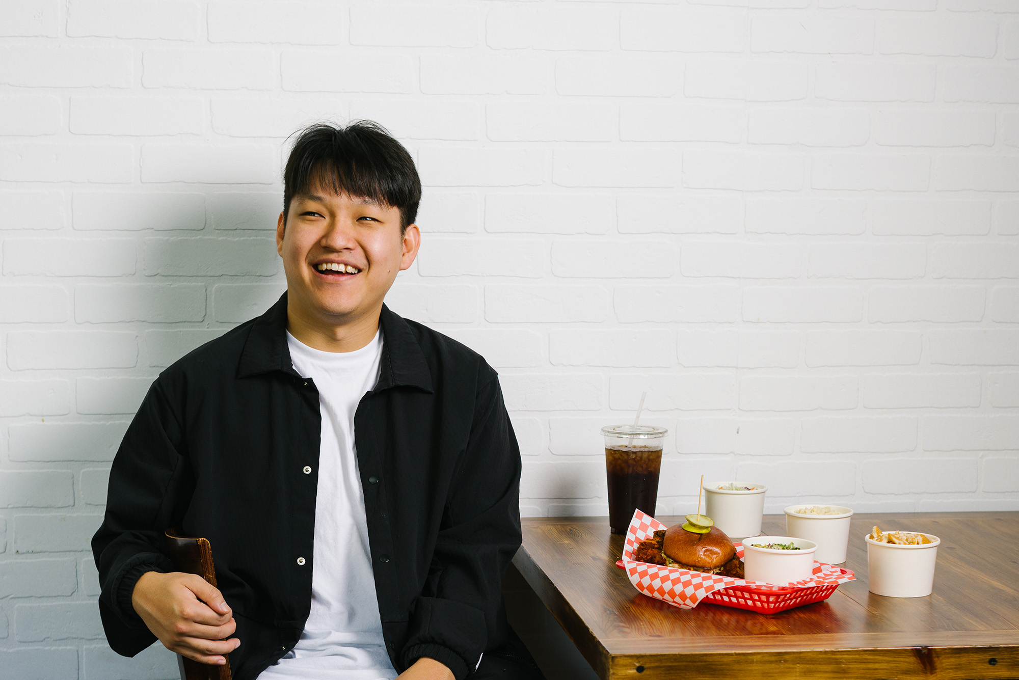 Photo: An Asian man with short black hair smiles as he sits in a chair next to a standard order at his chicken shop.