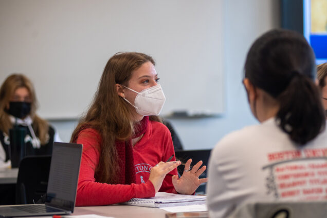 Photo: A young woman wearing a long-sleeved red shirt and a white KN95 mask, gesticulates as she talks in a classroom.