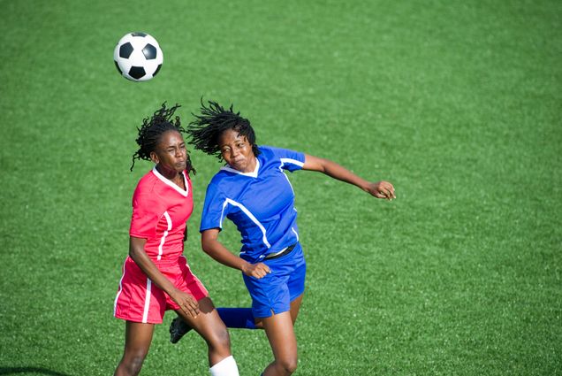 Photo: Two female soccer player, one in a red uniform and the other in blue, jump to head butt a ball flying through the air.