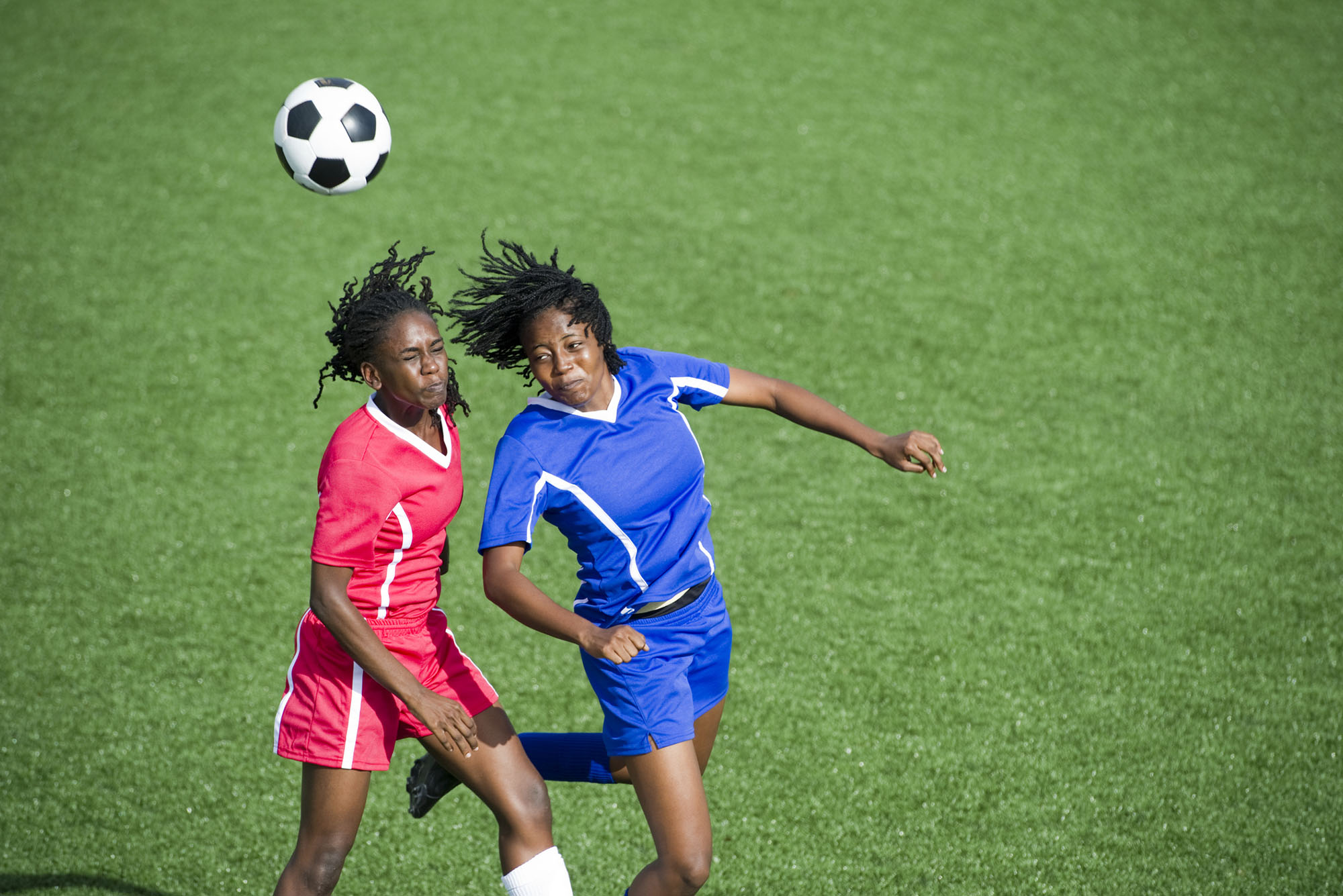 Photo: Two female soccer player, one in a red uniform and the other in blue, jump to head butt a ball flying through the air.