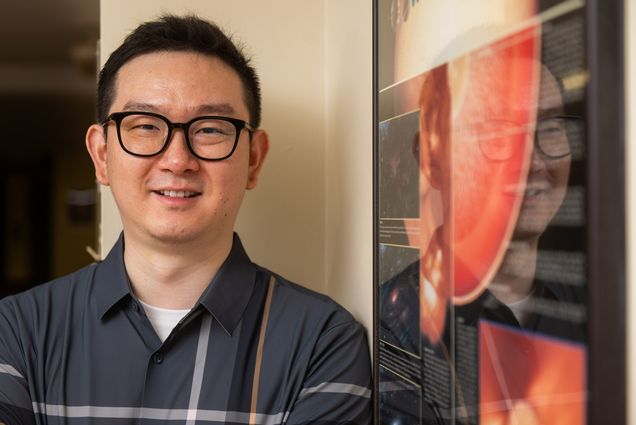 Photo: Chuanfei Dong, a Southeast Asian man wearing glasses and a dark blue-gray collared shirt, smiles and poses next to a a wall hanging explaining the workings of a planet.