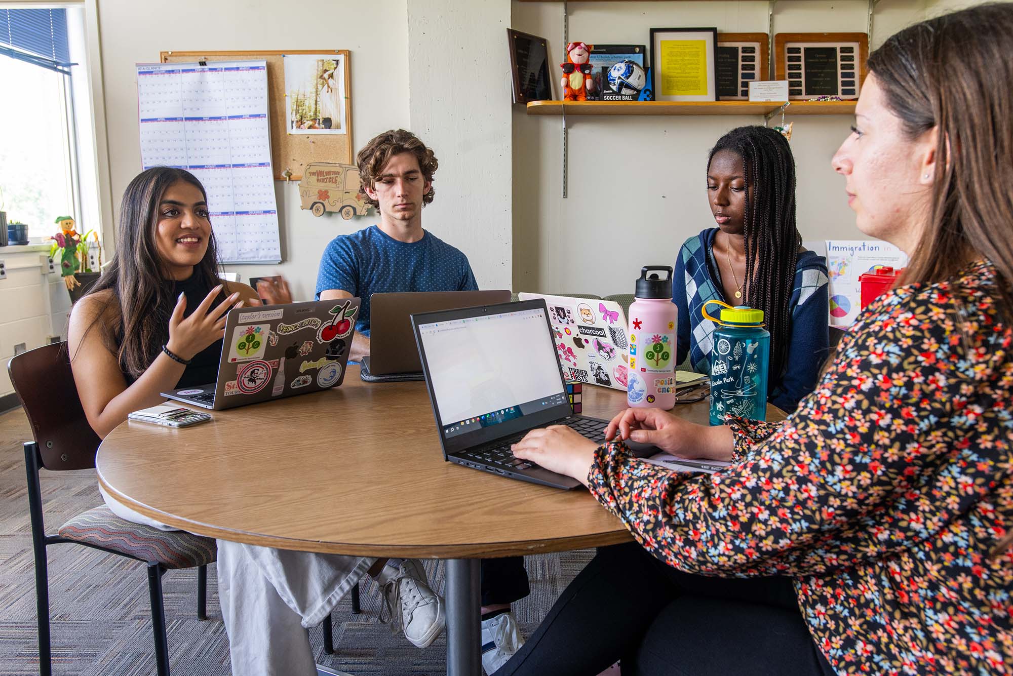 Photo: Four people, three college students and their advisor, sit at a round, wooden table. They work and look at individual laptop screens as they smile and collaborate.