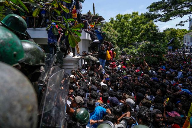 Photo: A massive group of protestors are shown storming Sri Lankan Prime Minister Ranil Wickremesinghe's office. The crowd piles and pushes at a large black metal gate and climb the white wall as officers in black riot gear try to fight them off.