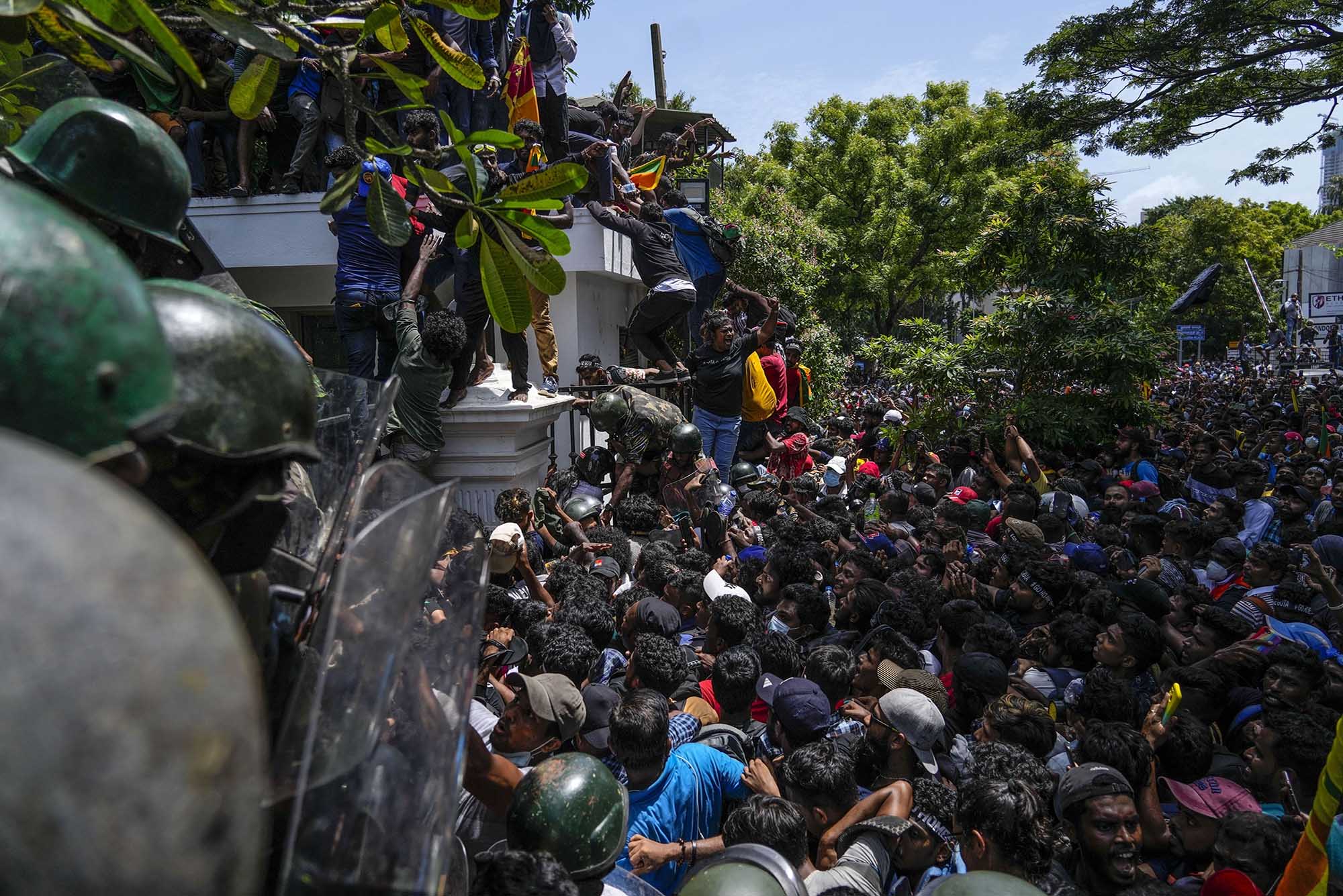 Photo: A massive group of protestors are shown storming Sri Lankan Prime Minister Ranil Wickremesinghe's office. The crowd piles and pushes at a large black metal gate and climb the white wall as officers in black riot gear try to fight them off.