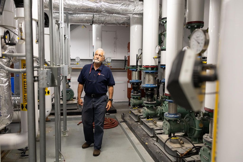 Photo: An older white man in an HVAC technician uniform stands in a room filled with pipes