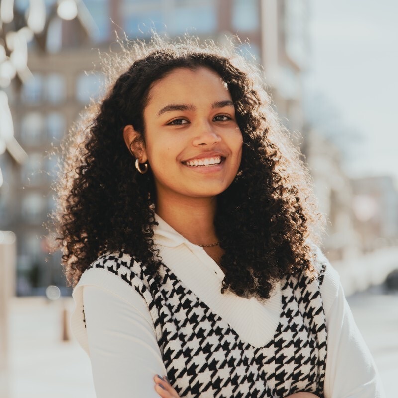 Photo: A headshot portrait of a young woman with long curly dark hair wearing a herringbone vest