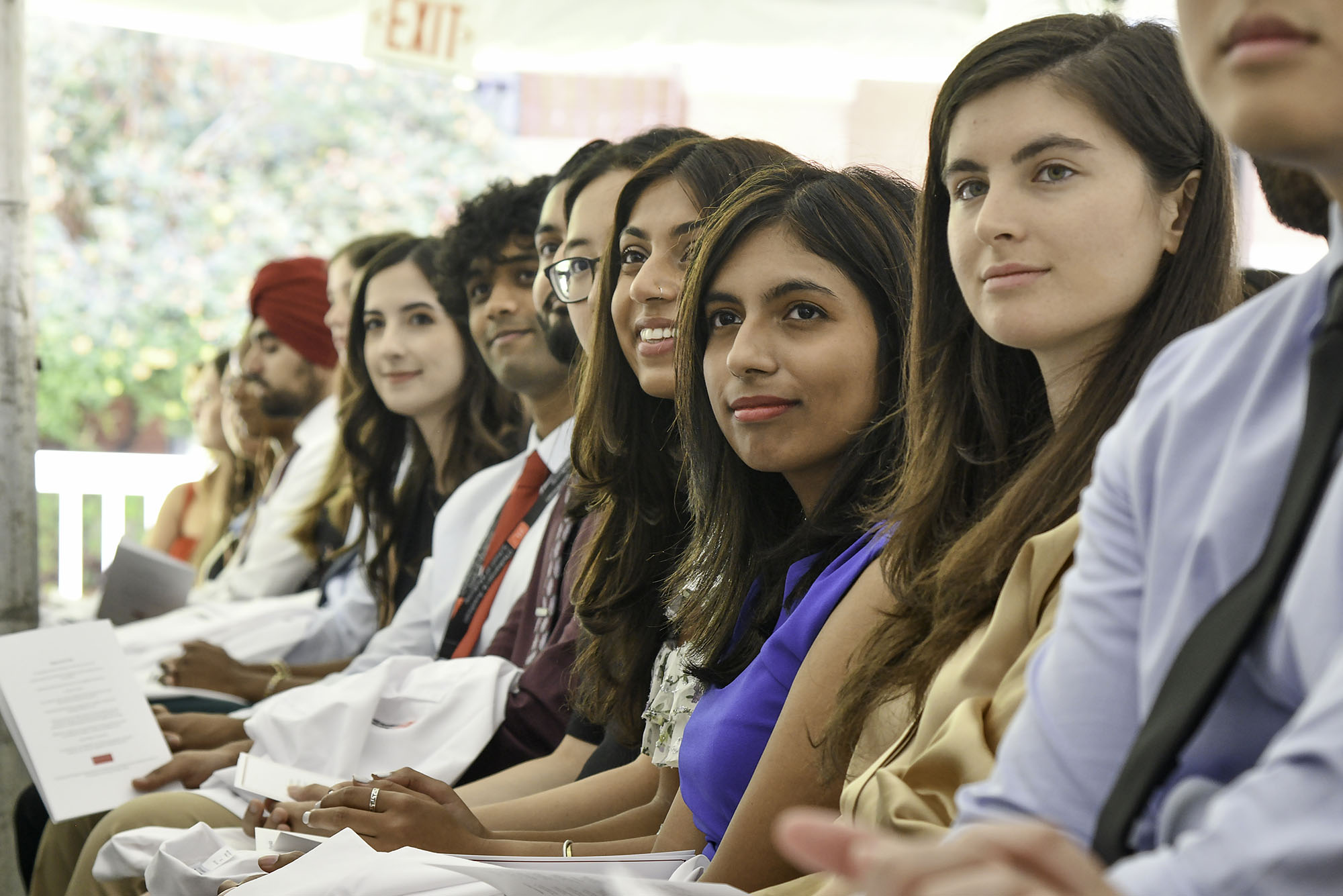 Photo: A lineup of students waiting to be called for their white coat ceremony. They all look to the left and up of the camera. The day is bright and sunny.
