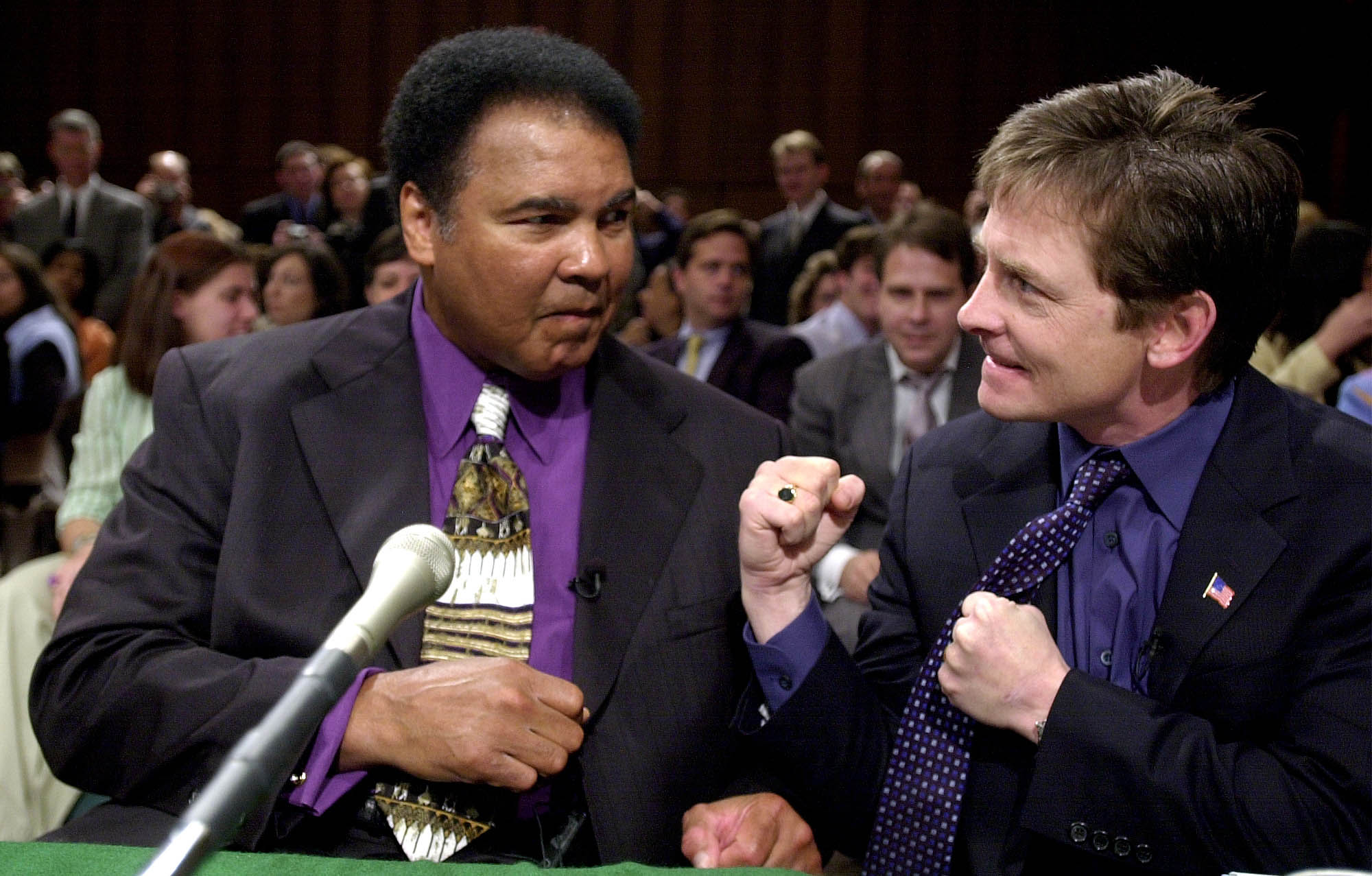 Photo: Boxer Muhammad Ali (left) and actor Michael J. Fox joke around. Ali, a Black man wearing a purple collared shirt, white and gold tie, and black blazer smiles as he jokingly raises his fist towards Fox, a white man wearing a dark purple collared shirt, purple tie, and black blazer. Fox laughs as he raises his own fists towards Ali in a joking way.