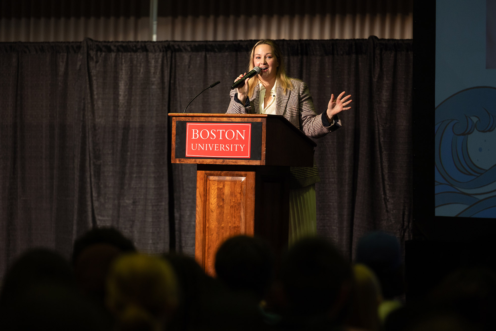 Photo: Boston City Councilor Sharon Durkan, a white woman with long blonde hair and wearing a white blouse and tweed blazer, speaks on stage into a mic. She stands behind a wooden podium with a red sign that reads "Boston University" and speaks to students in the audience.