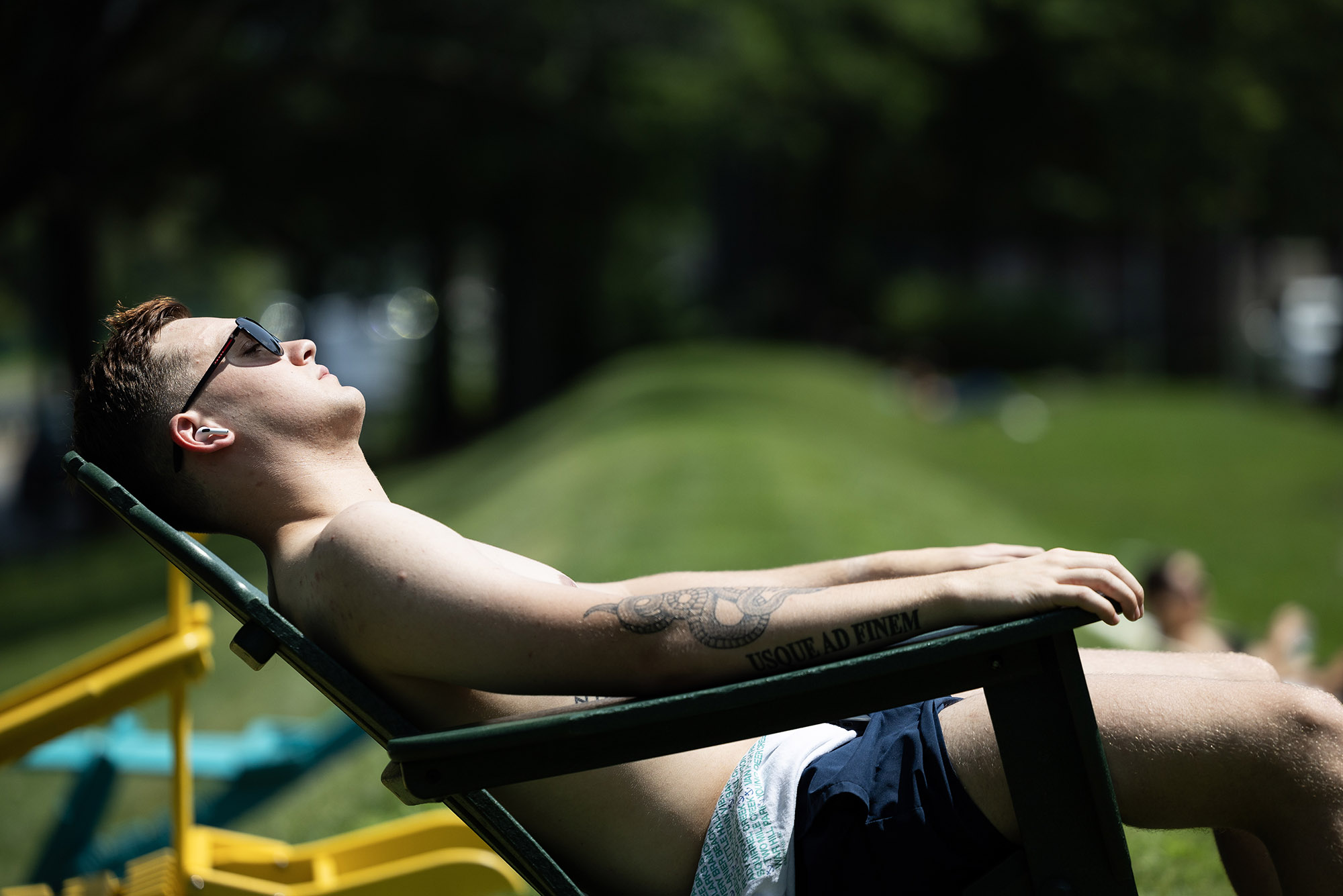 Photo: A young, white man lies out and relaxes in a beach chair. He is topless and wears swim trunks and sunglasses. The tattoo on his arm reads "Usque Ad Finem".