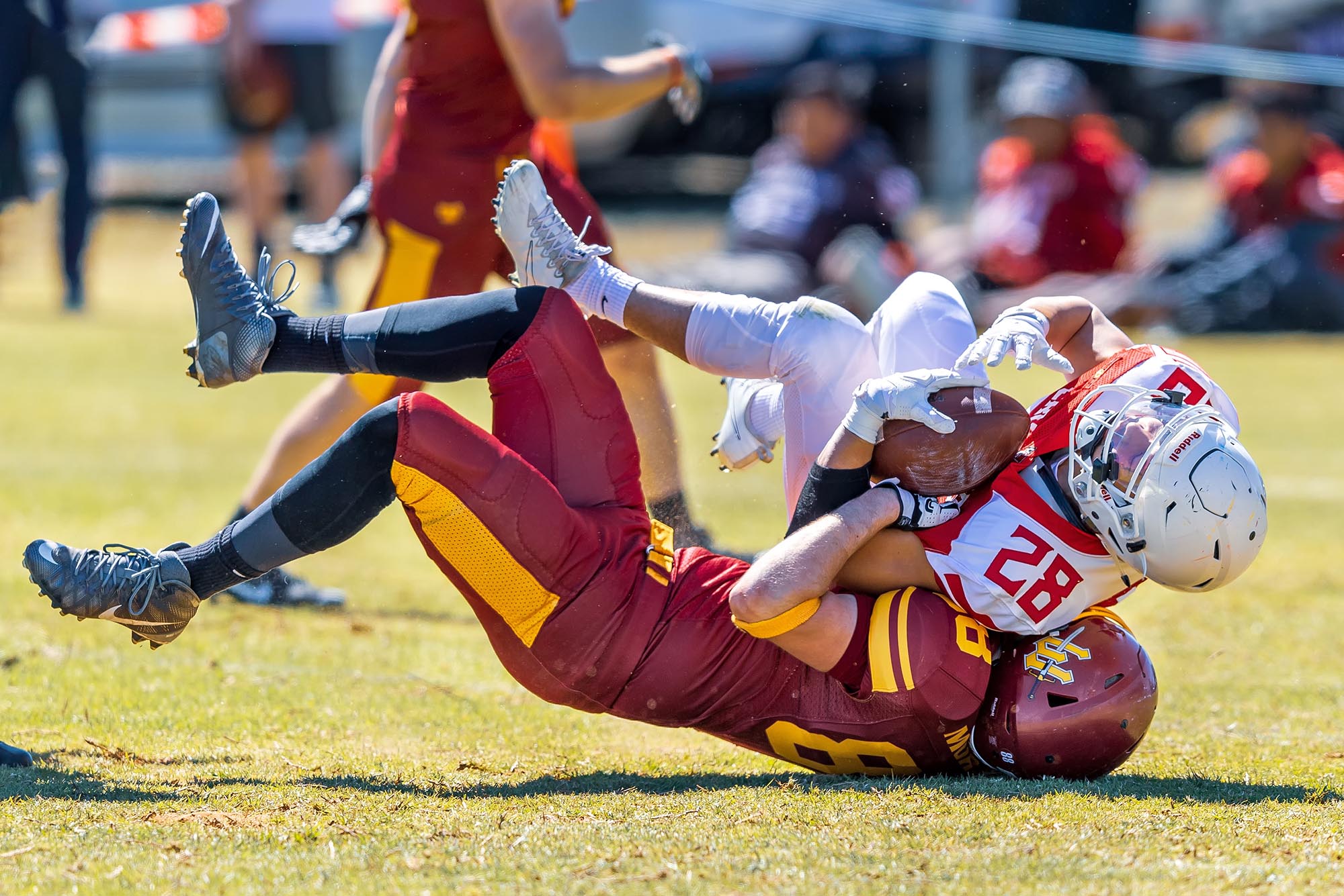 Photo: A person wearing a burgundy and yellow tackle football uniform and helmet is shown tackling a player in a white and red uniform and helmet tot he ground. The burgundy athlete has their head impacted to the ground as the white player falls on top of them.