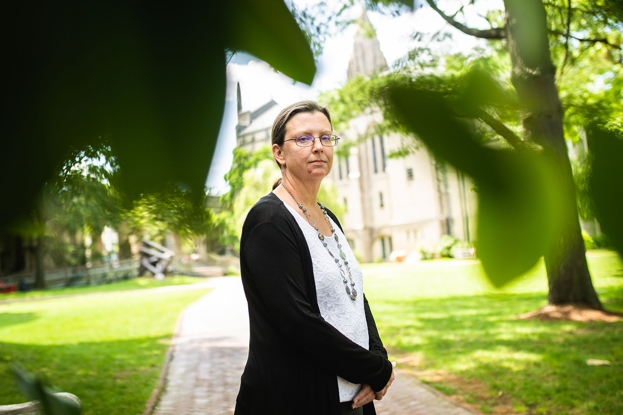 Photo: STH professor Rebecca Copeland poses for a photo on a bright green lawn. A white woman wearing glasses, a white blouse, black cardigan, and long beaded necklace, stands with hands placed in front of her. Foreground leaves frame her and an old church-like building is shown in the blurred background.
