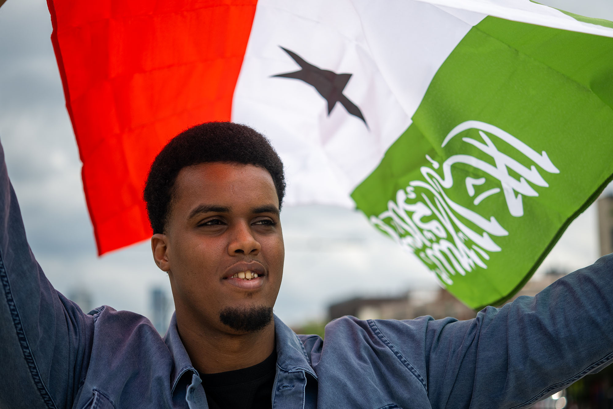Photo: Zakariya Hussein, a dark-skinned Somalian young man wearing a black shirt and blue collared lone-sleeved shirt smiles and holds the Somaliland flag above his head. The flag blows behind him, creating a striking portrait.