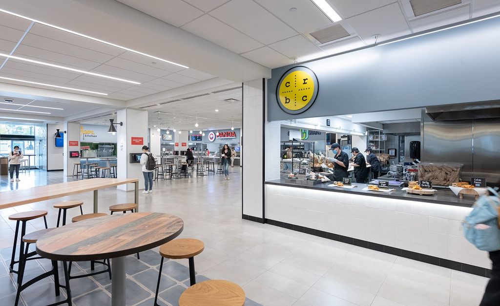 This photo was taken inside of Boston University's George Sherman (student) Union (GSU). The photo features a set of table and chairs with white and grey flooring. In the background, you can see students scattered around, either walking or sitting. On the left, there is a food window that has food displayed for grab.