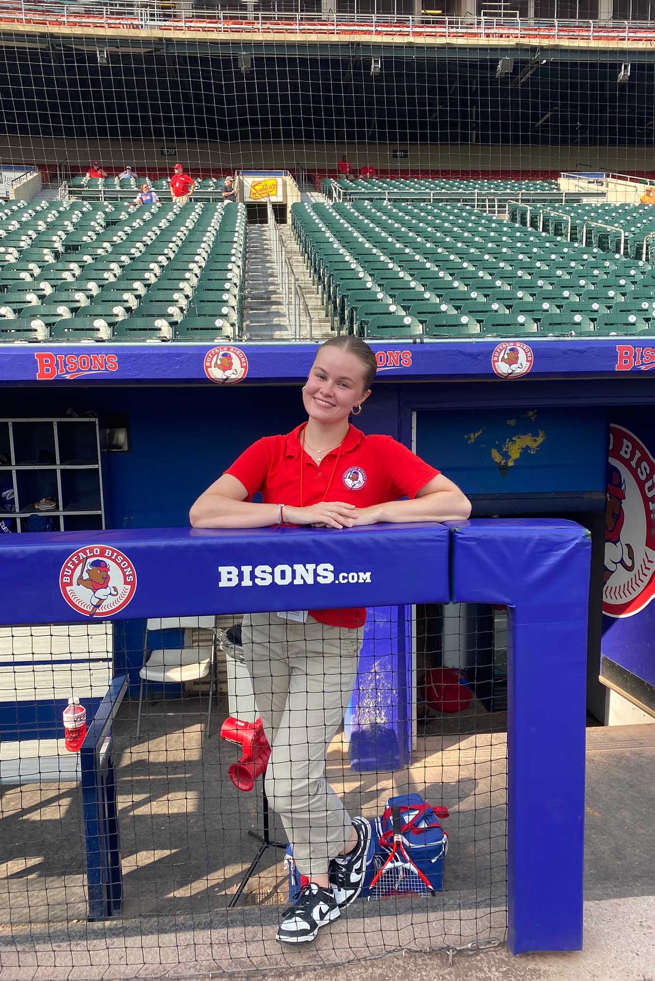 Photo: A young white woman with a red collared shirt and khakis leans on a blue fence in front of stadium seats. She smiles for the camera.