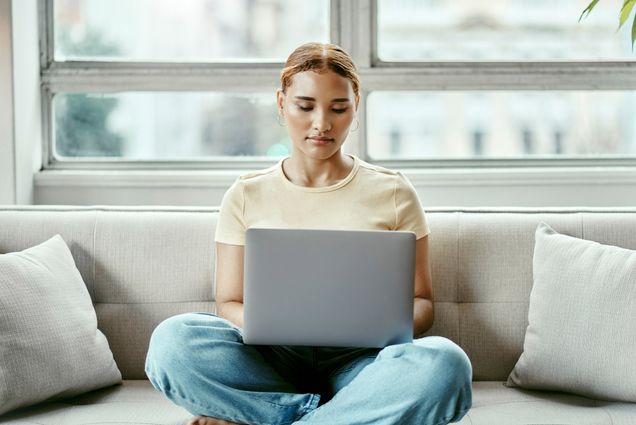 Photo: A young, tan woman with curly hair pulled back in a ponytail sits on a couch crossed-legged as she types on her silver laptop. She wears a yellow shirt and jeans as she sits in front of a bright window.