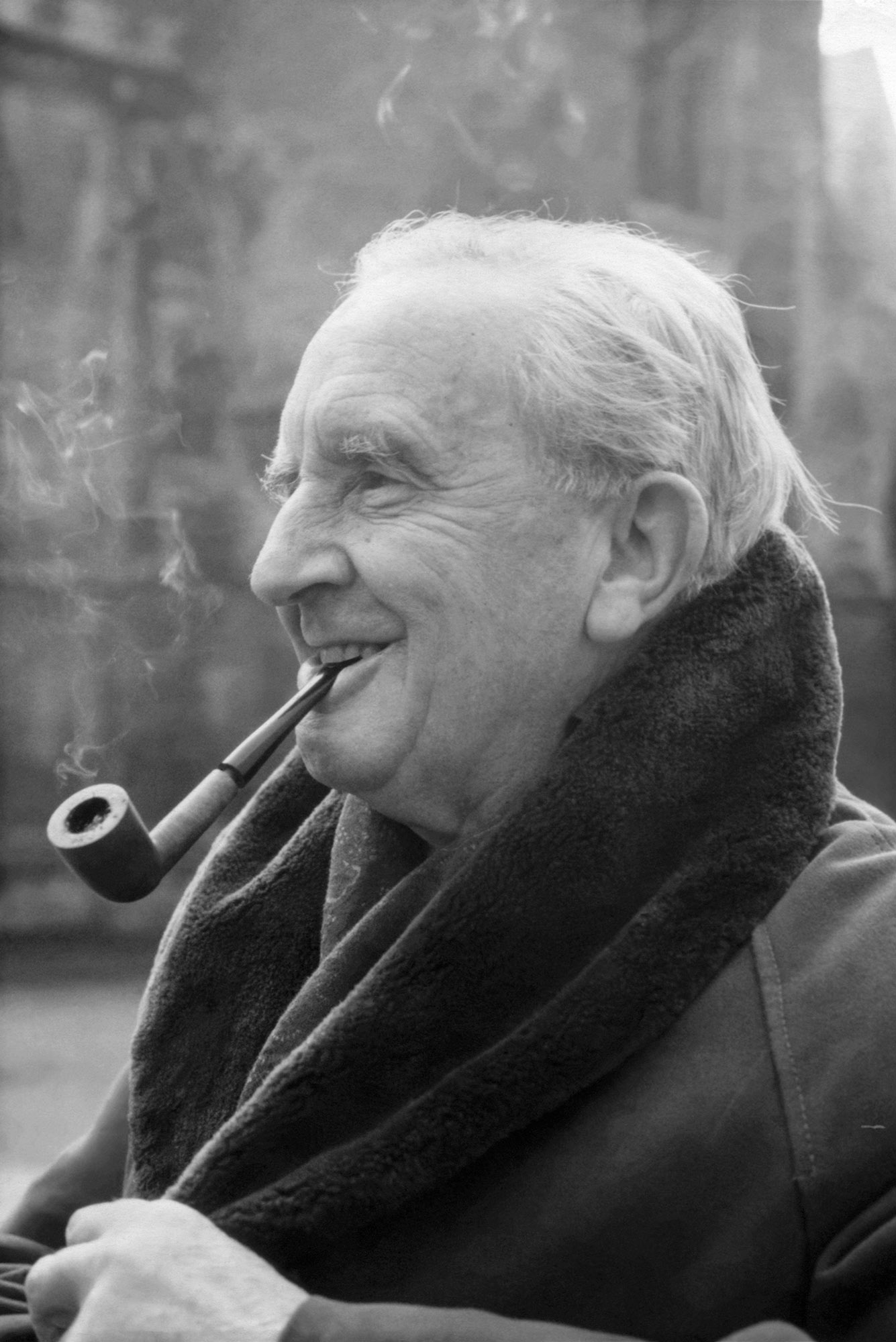 Photo: A black and white photo of J.R.R. Tolkien smoking a pipe. It hangs, leisurely, in his mouth and he smiles, amused, to something off camera. He wears a coat, fur-lined with an exposed and popped collar. His hair is short and white. The background is blurred, but he looks to be on a campus.