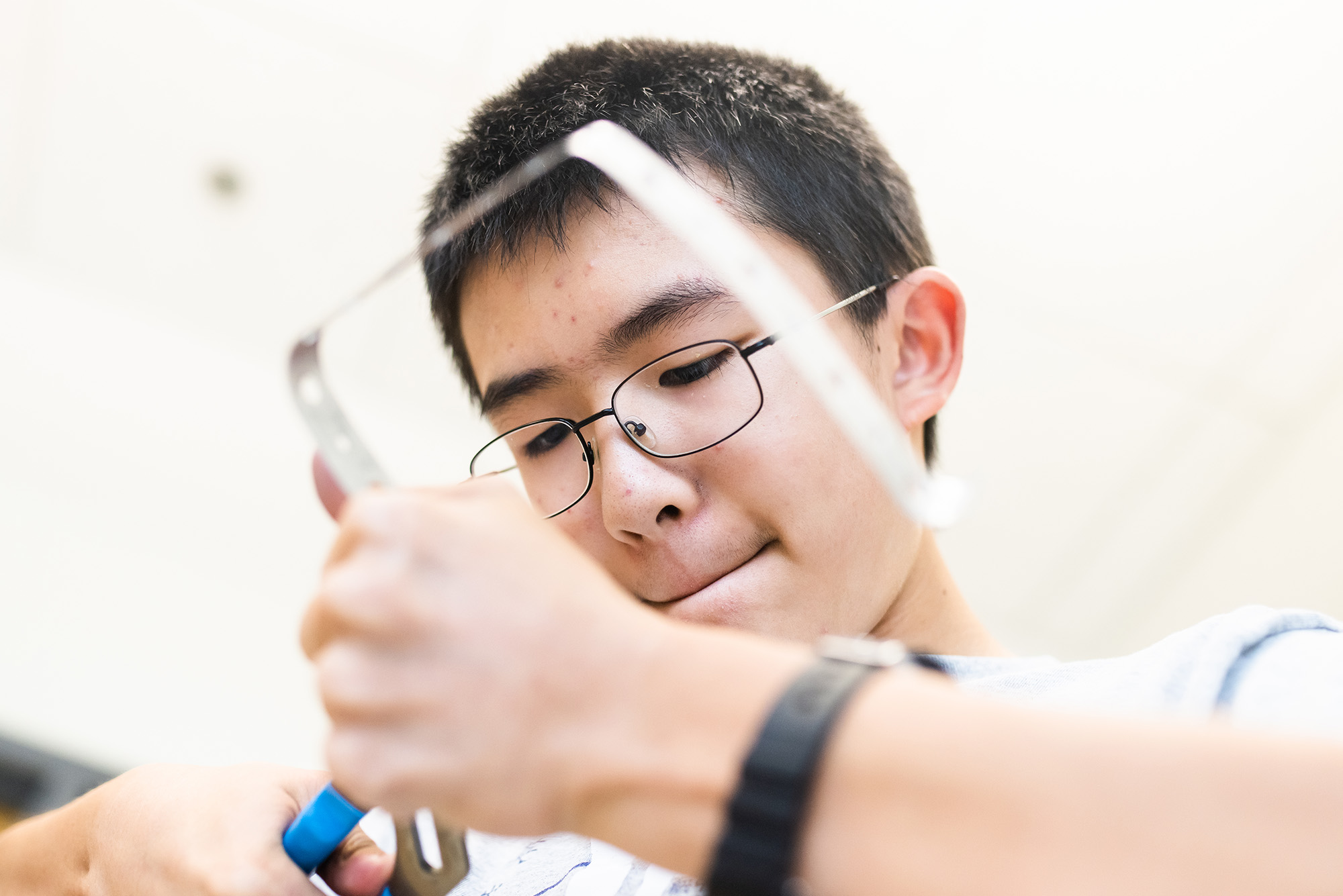 Photo: A Asian boy works on a metal strip that he is bending with pillars. He is looking down, concentrating on the object. He wears glasses and a black watch.