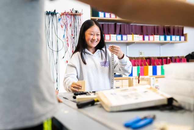 Photo: A young asian girl smiles and holds up a metal strip in a college-run program where she investigates more about STEM. She wears a gray hoodie.