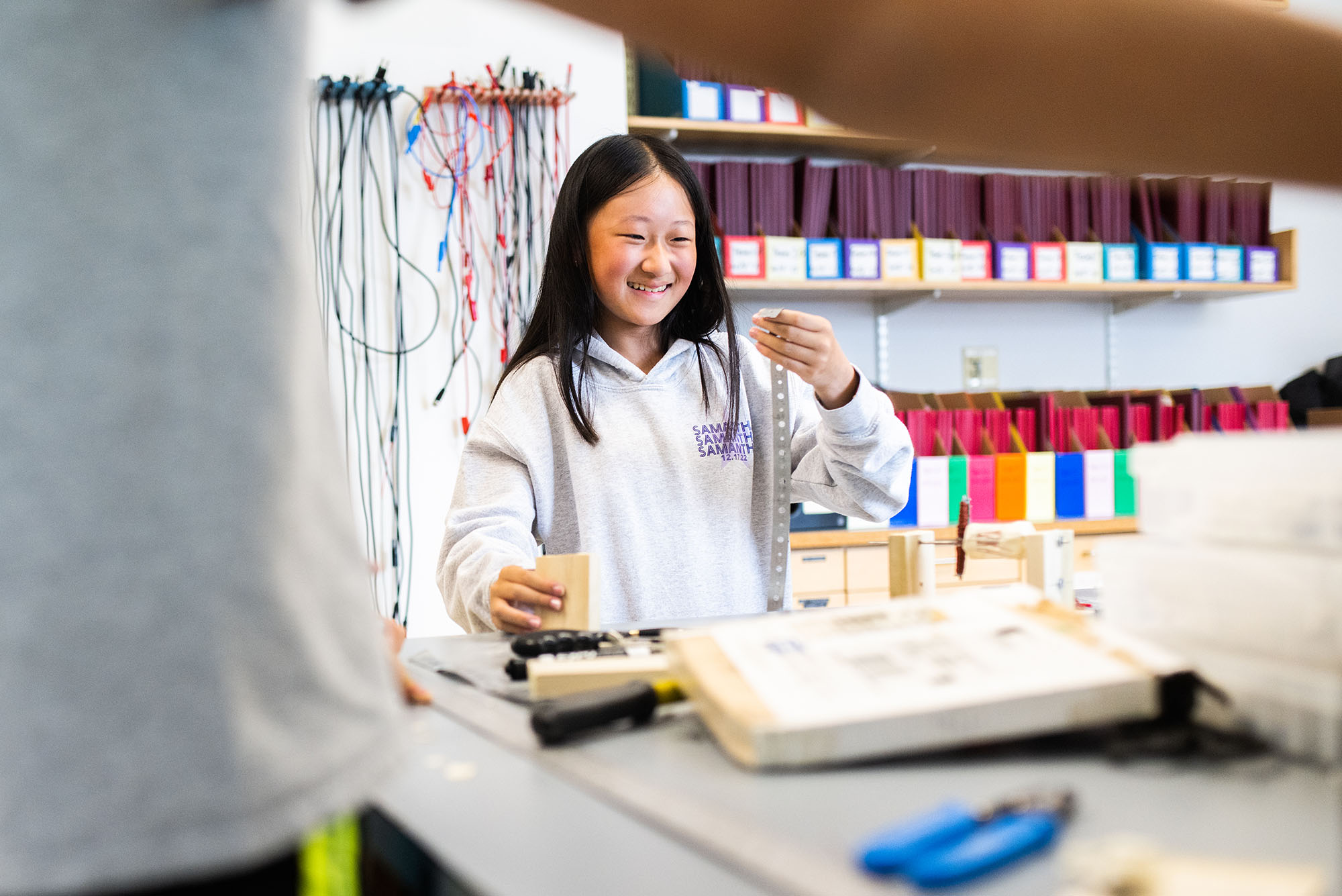 Photo: A young asian girl smiles and holds up a metal strip in a college-run program where she investigates more about STEM. She wears a gray hoodie.