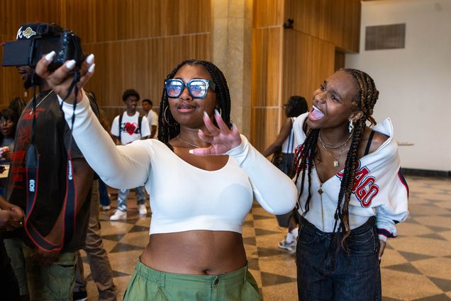 Photo: Two Black young women, one wearing a cropped long-sleeved white shirt, glasses, and olive green pants and the other wearing a grey sweatshirt and black pants, strike extravagant poses for a selfie photo.