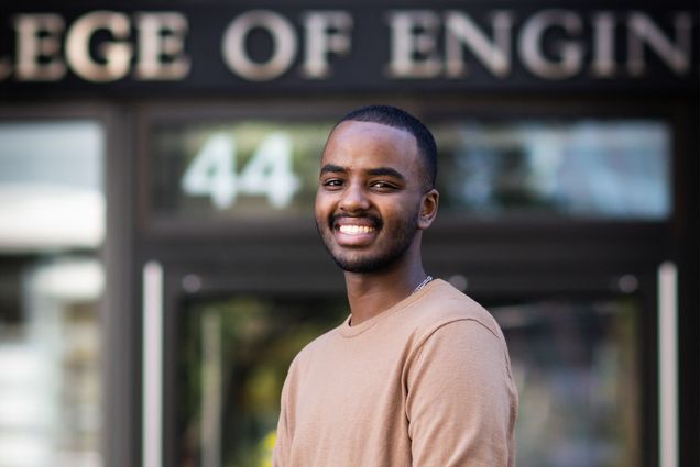 Photo: A young black man in a pink sweater stands with a smile and one hand in a pocket