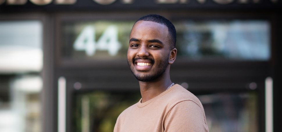 Photo: A young black man in a pink sweater stands with a smile and one hand in a pocket