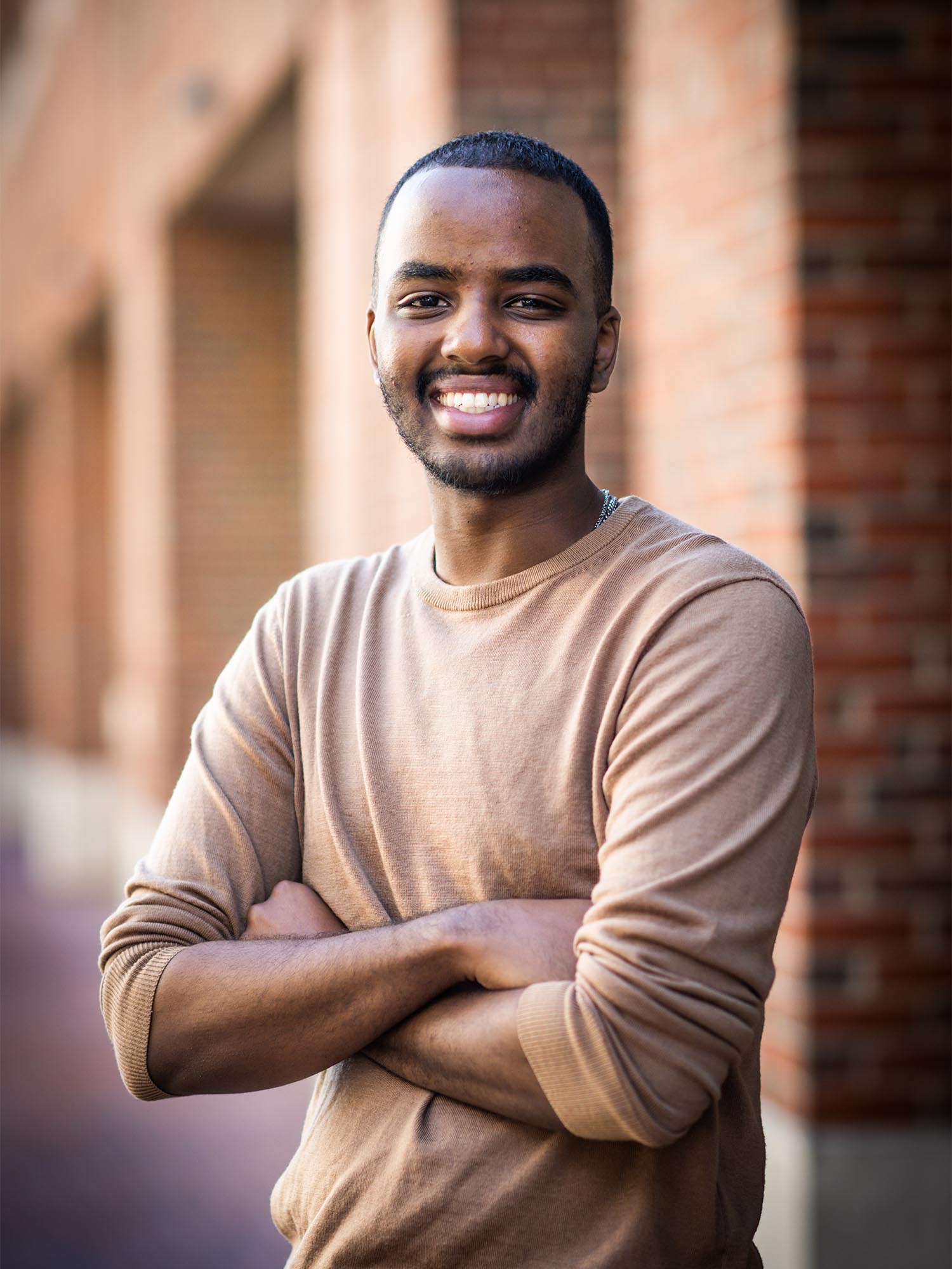Photo: A young black man in a pink sweater stands with a smile and arms crossed