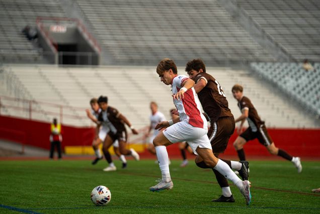 Photo: College soccer players race each other towards a soccer ball on a large green field in front of a row of metal bleachers