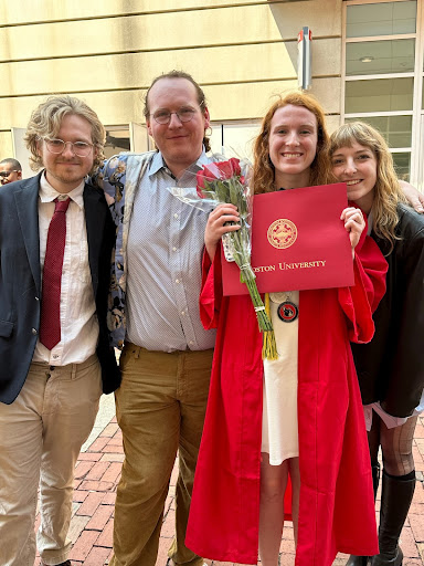This is a photo courtesy of Abigail Wood at Boston University's Commencement Ceremony from 2023. Wood is with three others. She has a white dress on, her red graduation gown, and red diploma from Boston University. She is holding roses in her hand. On her right, there is a blonde woman with a black jacket on leaning on her shoulder. On her left, there are two men. The man closest to her is in a blue button up shirt with tan pants and glasses on. The man next to him is wearing a suit jacket, a red tie, tan pants, and glasses. They both have light colored hair while the Abigail, the person in the graduation outfit, has red hair. They are all smiling in front of a tan colored building.
