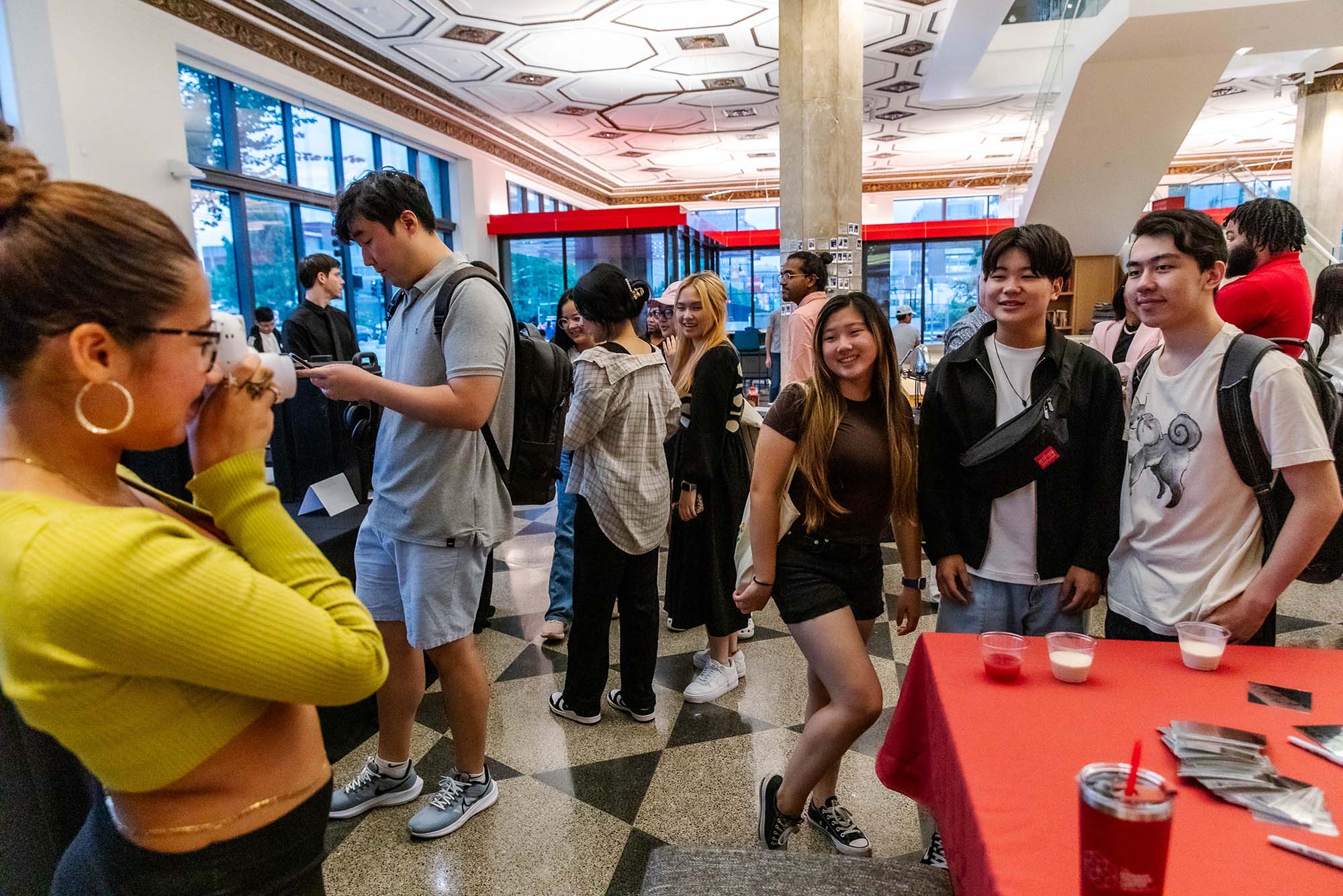 Photo: A student wearing a neon yellow long-sleeved top and jeans holds up a polaroid camera and takes a photo of a group of Asian students posing together to the right. Other students can be seen walking and mingling in the large reception room around and behind them.