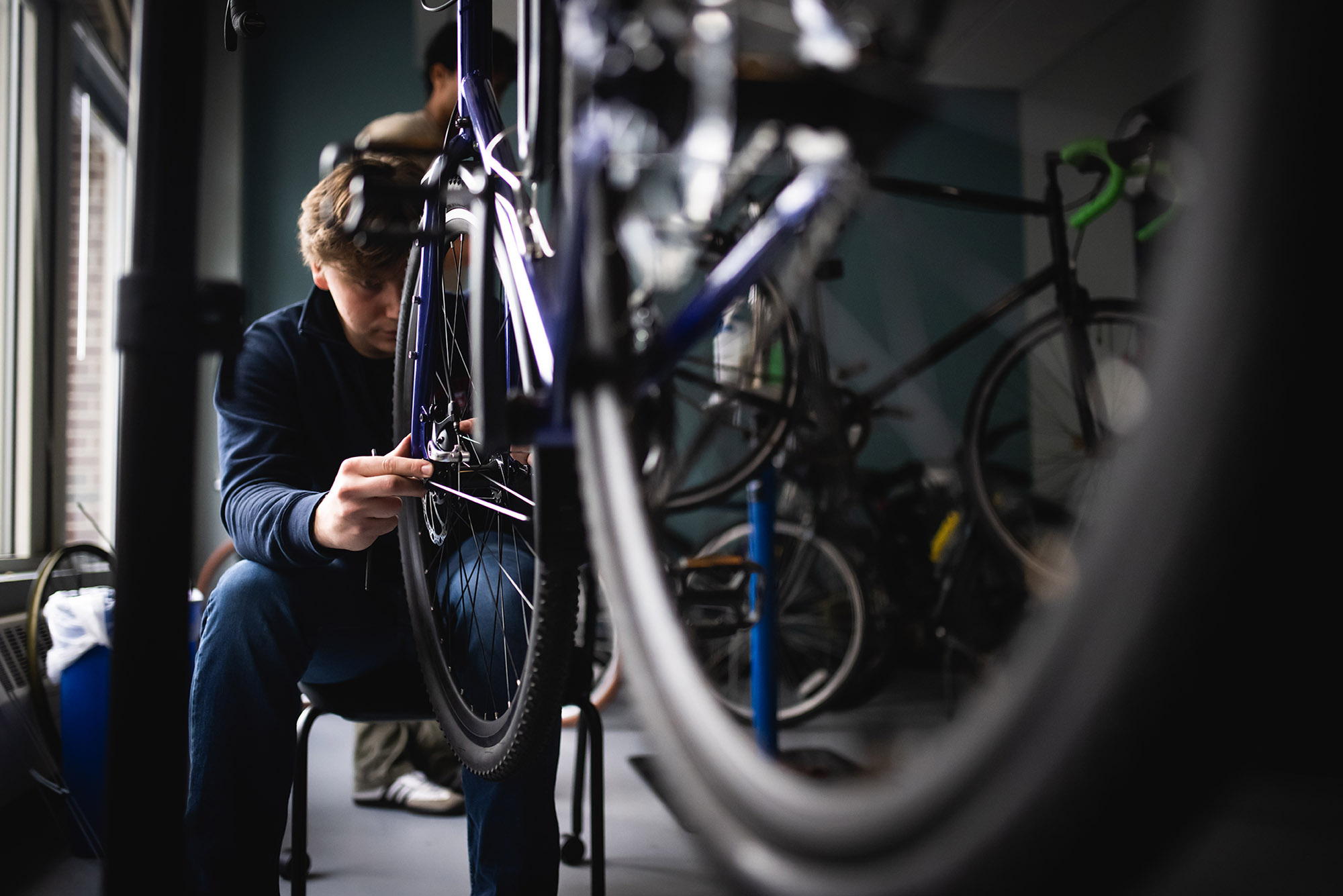 Photo: Close-up photo of a young man working on the front wheel of a mounted blue bicycle.