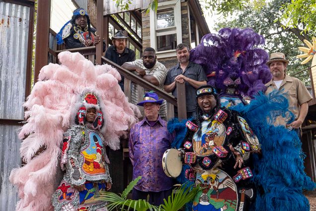 Photo: A group of people wearing bright, flamboyant Mardi Gras Indian outfits stand and pose in front of a large stoop. Large pink and purple feathers are showcased on outfits and headdresses.