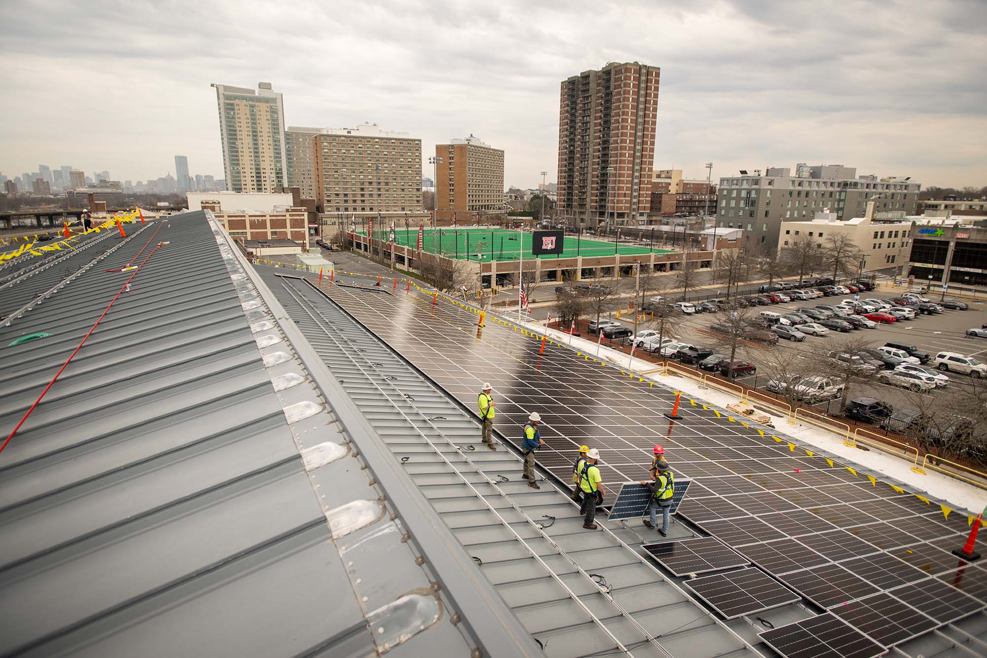 Photo: A team from Solect Energy instals what will ultimately be 1500 solar panels on the roof of the Track and Tennis Center April 6. A group of men wearing hard hats, neon vests, and construction gear work to place a large solar panel on the roof of a large building. In the distance the New Balance field, BU's West Campus, and various brick buildings can be seen.