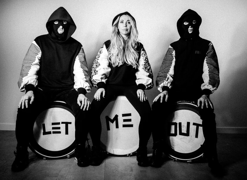 Photot: Black and white photo showing the Balaklava Blues, a trio sitting on 3 large drums. The front of the drums are spray-painted "let me out". The people on each end wear black hoods and full face masks while the person in the middle sits unmasked wearing a letterman jacket.
