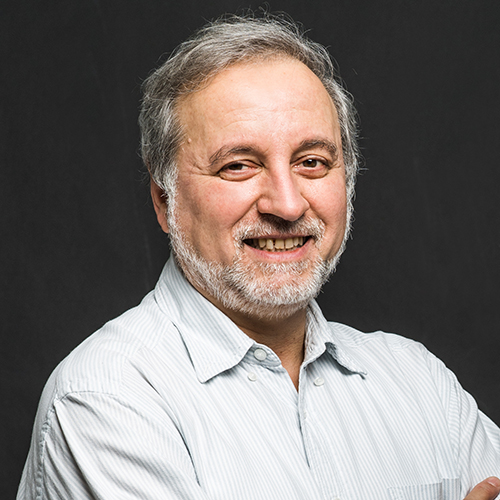 Photo: Bela Suki, an older white man with silver grey hair and beard and wearing a light blue collared shirt, smiles and poses with arms crossed in front of a dark background.