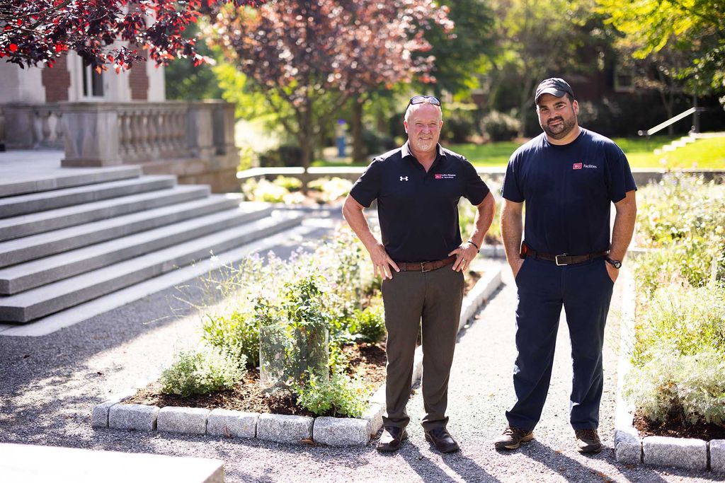 Photo: Greg Limerick, manager of trucking and grounds, left, and Matt Pereira, groundsworker, right, work at the CPO's new pollinator garden at Hawes Street on Wednesday. Two men wearing navy blue shirts and grey pants stand and pose in front of a blooming pollinator garden on BU's campus.