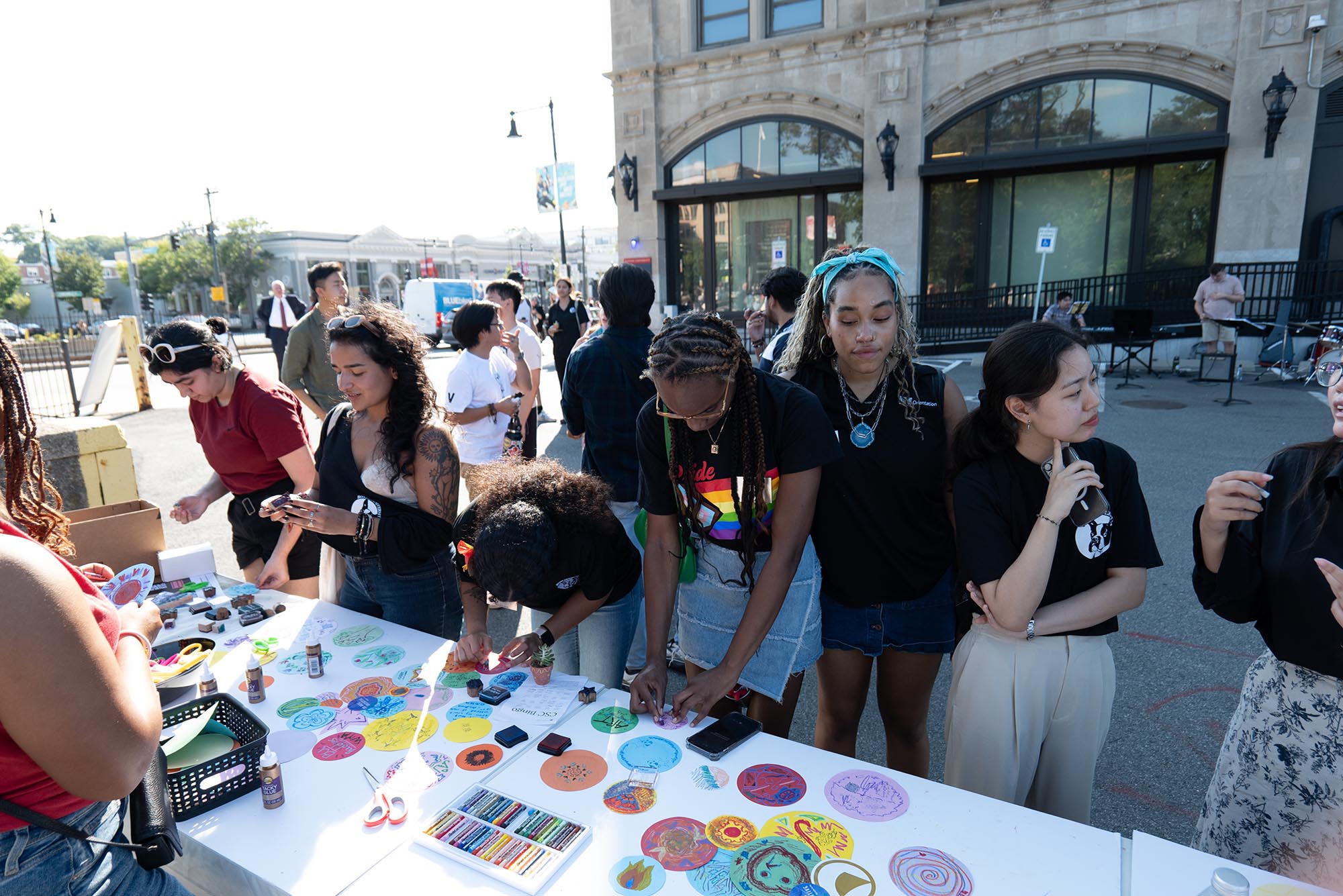 Photo: Students line up on a street at booths featuring free stickers, gear, and knick knacks as they laugh and smile on a sunny day.