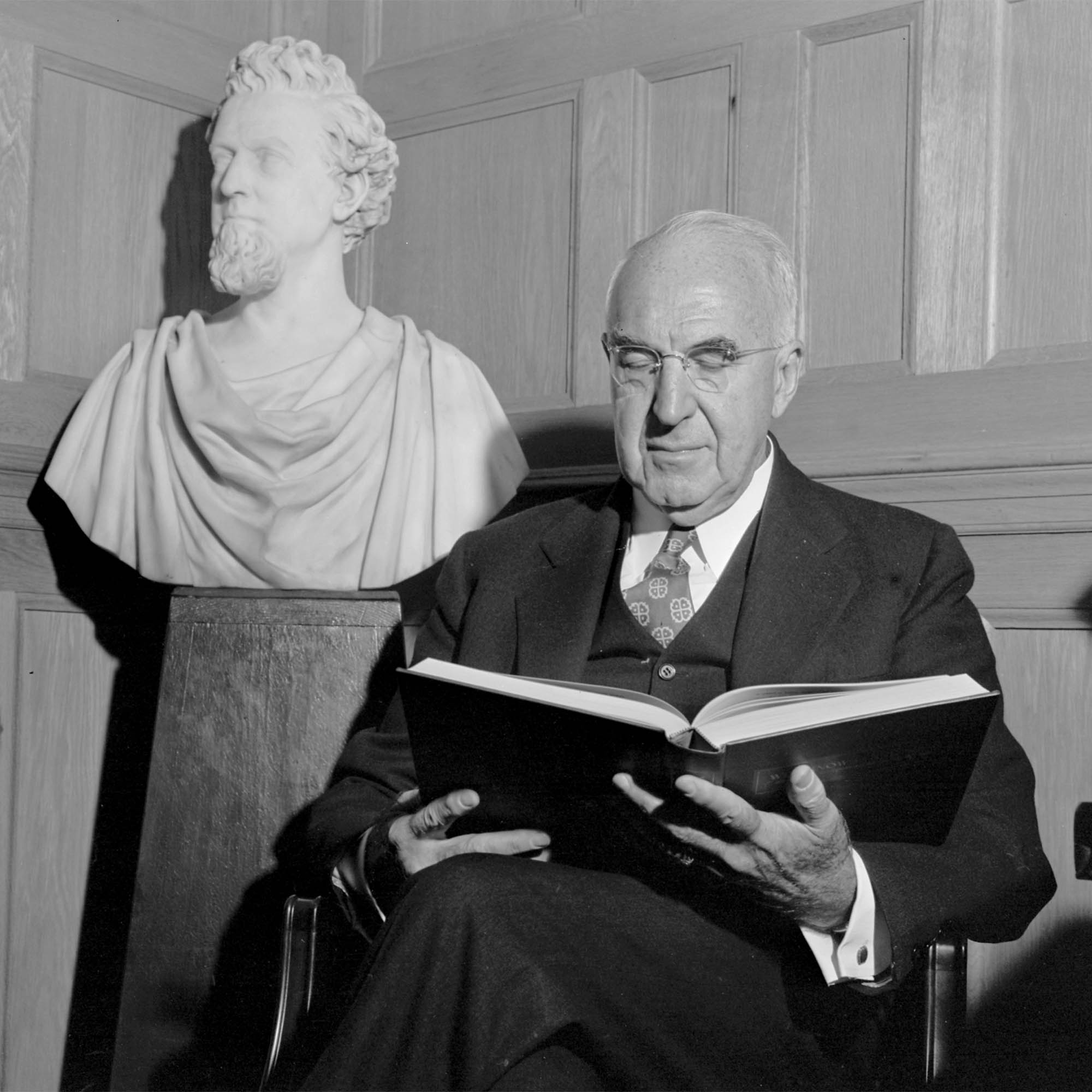 Photo: A man in a suit reading a book sits beside a greek bust of a presumed philosopher