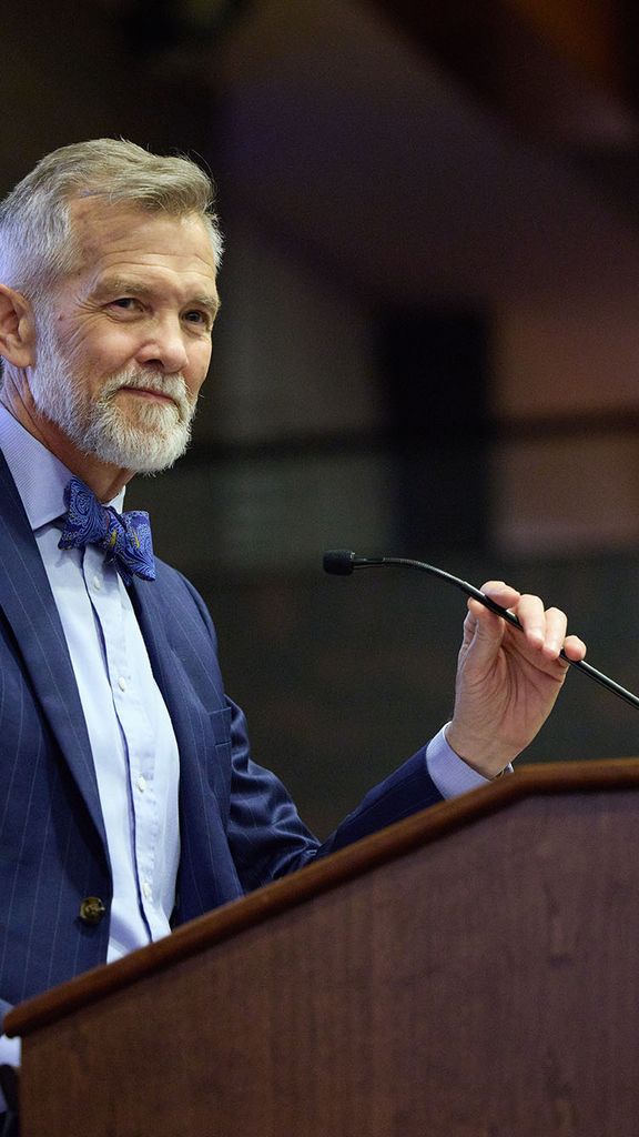 Photo: David Chard, a white man with short gray hair and short gray beard. He wears a blue suit and maroon tie, smiles and stands at a wooden podium with a red "Boston University" sign on it. He holds a small microphone in one hand as he looks to an unseen audience.