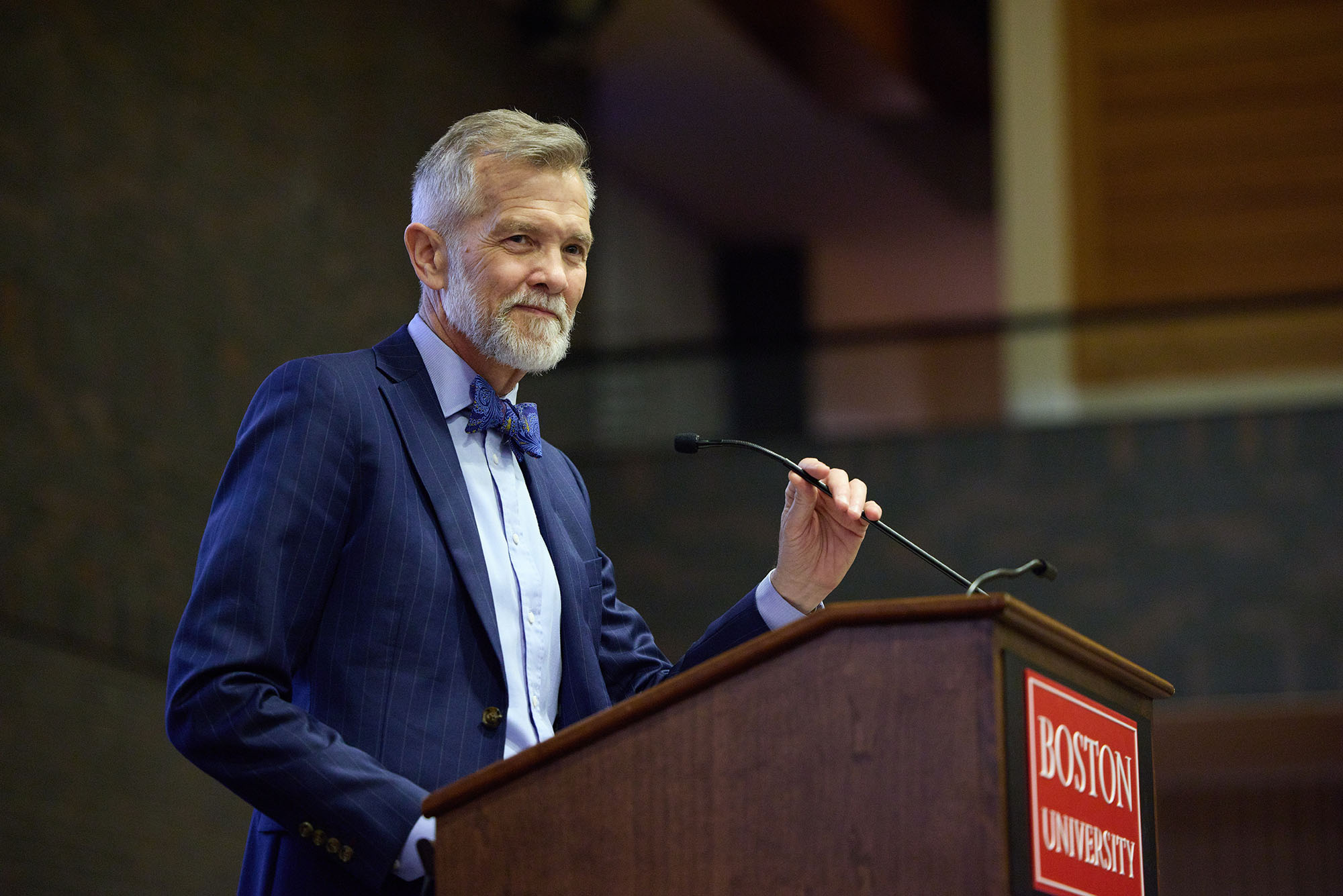 Photo: David Chard, a white man with short gray hair and short gray beard. He wears a blue suit and maroon tie, smiles and stands at a wooden podium with a red "Boston University" sign on it. He holds a small microphone in one hand as he looks to an unseen audience.